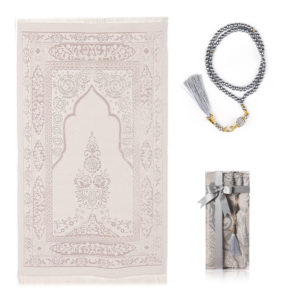 Un tapis de prière gris est étendu sur un fond blanc. Un chapelet assorti est à sa droite et une petite boîte également.