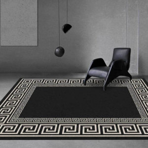 Un tapis noir avec des formes géométriques beige est installé au sol dans un salon très moderne. Les murs sont en béton gris, un cadre uni est sur le mur. Il est a une lampe noir suspendue et une chaise design complètement noire.