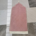 Un tapis rose épais est posé sur un carrelage. C'est un tapis épais et doux avec des franges dorées en bas et un pompon en haut. Il a une forme de porte orientale. Le coeur du tapis est orné de motifs orientaux.