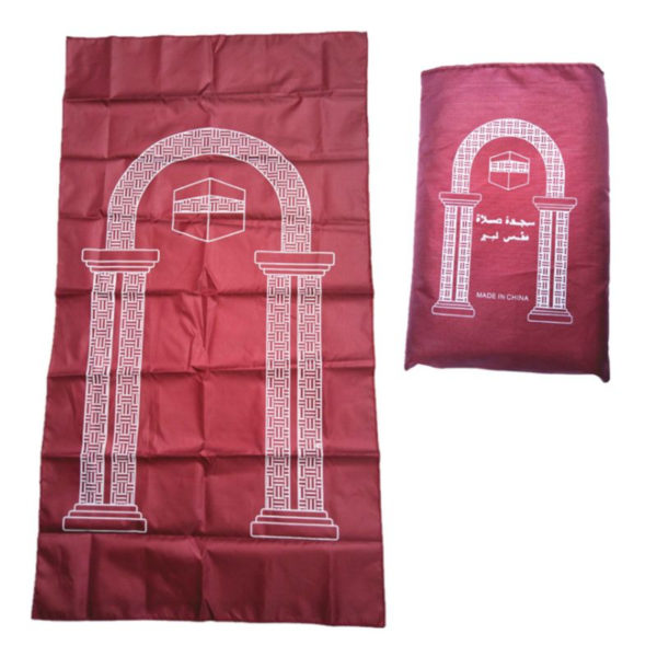 Un tapis rouge décoré d'une arche est de la kaaba sur un fond blanc. Le tissu est imperméable et une pochette de transport est posée à côté.