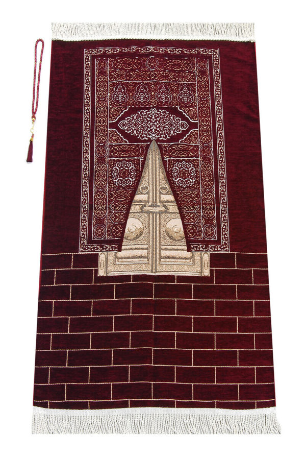 Un tapis rouge est étendu sur un fond blanc. Il est décoré avec le motif des portes dorées de la Kaaba et de franges en haut et en bas. Il est accompagné d'un chapelet de prière.