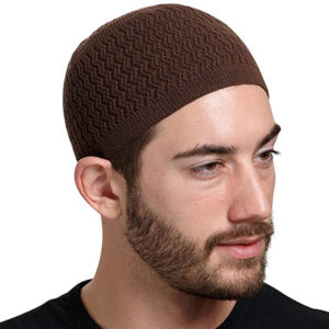 Un homme se tient de profil sur un fond blanc. Il porte une barbe un kufi tricoté marron sur la tête.