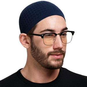 Un homme à barbe avec des lunettes porte un kufi bleu tricoté sur la tête sur un fond blanc.