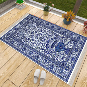 Tapis de prière bleu à motifs persan. Un tapis pais et anti-dérapant. Le tapis est sur du parquet, une paire de chaussons est pos à côté ainsi que des pots de fleurs.