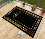 Un tapis de prière noir et doré posé sur du parquet à côté d'une fenêtre. Des chaussons sont posés à côté du tapis ainsi que des pots de fleurs. Le tapis est majoritairement noir avec des dorures sur son contour et un motif de porte doré au milieu.