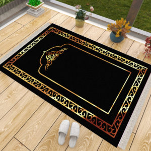 Un tapis de prière noir et doré posé sur du parquet à côté d'une fenêtre. Des chaussons sont posés à côté du tapis ainsi que des pots de fleurs. Le tapis est majoritairement noir avec des dorures sur son contour et un motif de porte doré au milieu.