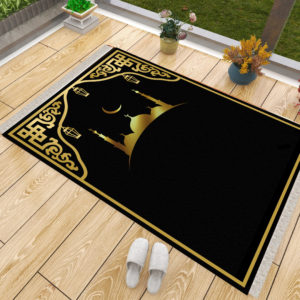 Un tapis de prière noir et doré posé sur du parquet à côté d'une fenêtre. Des chaussons sont posés à côté du tapis ainsi que des pots de fleurs. Le tapis est majoritairement noir et présente des dorures sur les coins supérieur et une mosquée avec un croissant de lune au dessus.