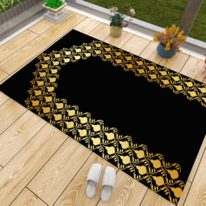 Un tapis de prière noir et doré posé sur du parquet à côté d'une fenêtre. Des chaussons sont posés à côté du tapis ainsi que des pots de fleurs. Le tapis est majoritairement noir avec une dorure en forme de porte.