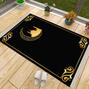 Un tapis de prière noir et doré posé sur du parquet à côté d'une fenêtre. Des chaussons sont posés à côté du tapis ainsi que des pots de fleurs. Le tapis est majoritairement noir et présente des dorures dans les coins et une mosquée dans un croissant de lune.