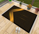 Un tapis de prière noir et doré posé sur du parquet à côté d'une fenêtre. Des chaussons sont posés à côté du tapis ainsi que des pots de fleurs. Le tapis est majoritairement noir avec un motif doré comme un coin, celui de la kaaba.