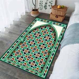 Superbe tapis de prière à dominante verte, aux motifs mosaïques marron, verts et beiges, de grande taille. Il se trouve dans une chambre avec un lit aux draps blancs, à côté duquel se trouve des boîtes en rotin