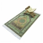 Tapis de prière en coton vert avec des motifs à fleurs. Sur le tapis un tabouret est posé avec un livre du coran dessus.