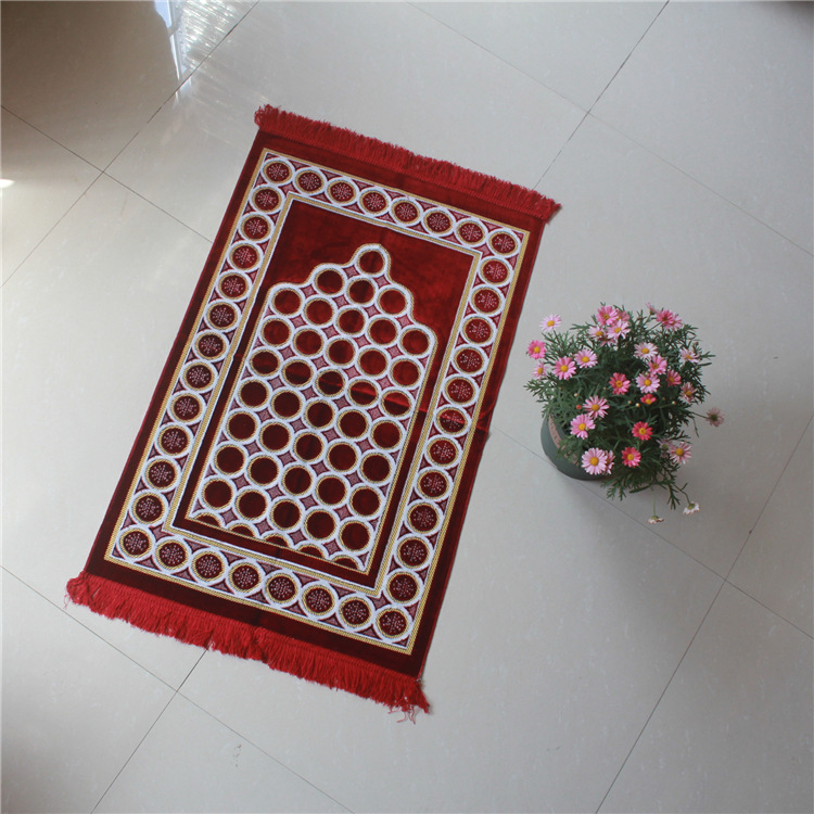 Tapis de prière rouge en velours avec des motifs rond. LE tapis est posé sur le sol à côté d'un pot de fleur.