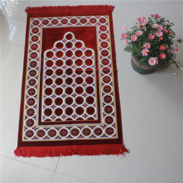 Tapis de prière rouge en velours avec des motifs rond. LE tapis est posé sur le sol à côté d'un pot de fleur.