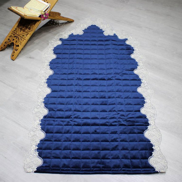 Tapis de prière bleu molletonné avec un contour en dentelle. Le tapis est posé sur le sol avec un coran ouvert sur un chevalet à cöté.