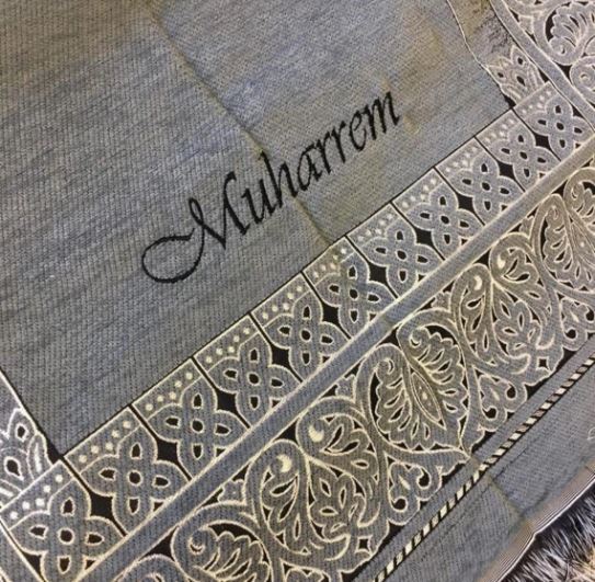 Un tapis de prière gris est orné de broderies délicates. Le prénom Muharrem est brodé sur le tapis de prière.