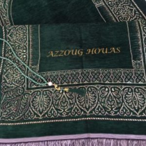 Un tapis de prière vert est posé plié sur le sol avec un chapelet. Il est décoré de motifs orientaux et de franges et un nom est brodé en fil doré.