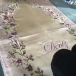 Un tapis beige doré satiné est étendu sur le sol. Il est décoré de franges et de fleurs violettes et de feuillage.