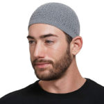 Un homme à barbe se tient de 3/4 profil devant un fond blanc. Il porte un pull noir et un kufi gris tricoté sur la tête.