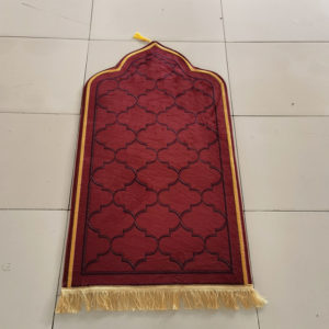 Un tapis à forme rectangulaire avec le bout en pointe est posé sur du carrelage. Il a des franges dorés et un pompon à l'extrémité. Il est rouge avec les bords dorés.