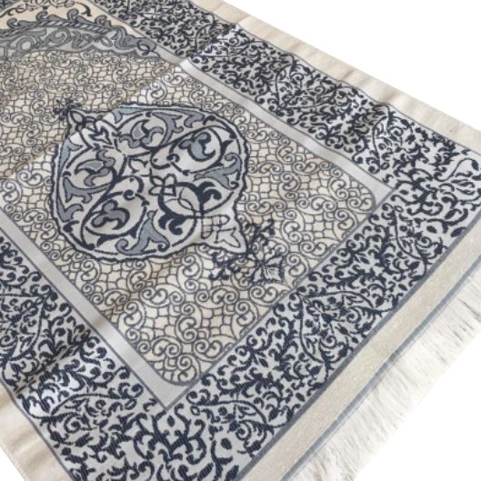 Un tapis de prière gris est étendu sur un fond blanc. Il est décoré de motifs orientaux et de franges.