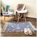 Ce tapis coloré est rectangulaire et avec des motifs cachemire. Ses bords ont des franges. Il est placé au pied d'une chaise et d'une petite table. Des chaussons sont disposés dessus.