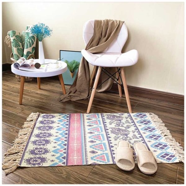 Ce tapis coloré est rectangulaire et avec des motifs losanges et fleurs. Ses bords ont des franges. Il est placé au pied d'une chaise et d'une petite table. Des chaussons sont disposés dessus..