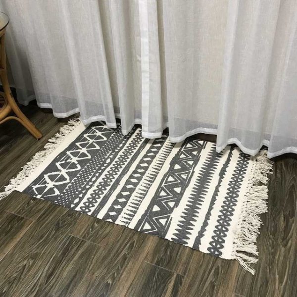 Ce tapis blanc et noir est rectangulaire et avec des motifs losanges. Ses bords ont des franges. Il est placé au pied d'un rideau blanc transparent.