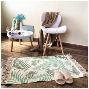 Ce tapis coloré est rectangulaire et avec des motifs feuillages. Ses bords ont des franges. Il est placé au pied d'une chaise et d'une petite table. Des chaussons sont disposés dessus.