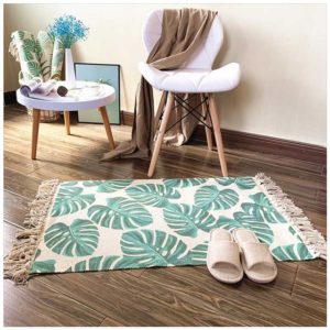 Ce tapis coloré est rectangulaire à motif plante exotique. Ses bords ont des franges. Il est placé au pied d'une chaise et d'une petite table. Des chaussons sont disposés dessus.