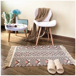 Ce tapis coloré est rectangulaire à motifs incas. Ses bords ont des franges. Il est placé au pied d'une chaise et d'une petite table. Des chaussons sont disposés dessus.