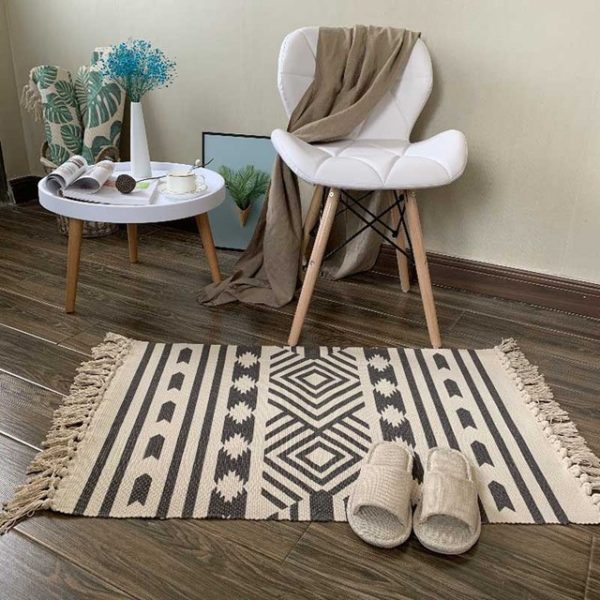 Ce tapis blanc et noir est rectangulaire et avec des motifs losanges. Ses bords ont des franges. Il est placé au pied d'une chaise et d'une petite table. Des chaussons sont disposés dessus.