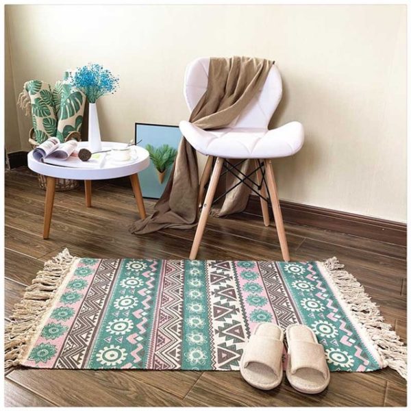 Ce tapis coloré est rectangulaire et avec des motifs roses et verts. Ses bords ont des franges. Il est placé au pied d'une chaise et d'une petite table. Des chaussons sont disposés dessus.