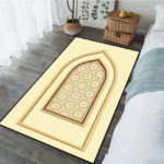 Tapis de prière beige avec un motifs de porte orientale. Le tapis st posé dans une chambre entre un lit et une fenêtre.