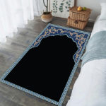 Tapis de prière noir avec un contour bleu. Le tapis est posé sur du parquet dans une chambre entre un lit et une fenêtre.