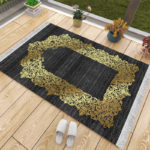 Un tapis de prière noir et doré posé sur du parquet à côté d'une fenêtre. Des chaussons sont posés à côté du tapis ainsi que des pots de fleurs. Le tapis est majoritairement noir avec une magnifique dorure en forme de porte.