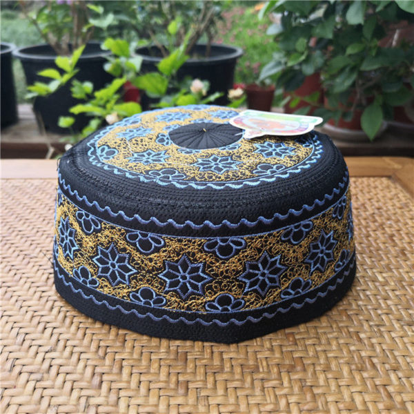 Un kufi est posé sur une table en bois en extérieur. Il est noir et décoré de fil bleu et doré. Les broderies forment des motifs géométriques et floraux sur tout le kufi.